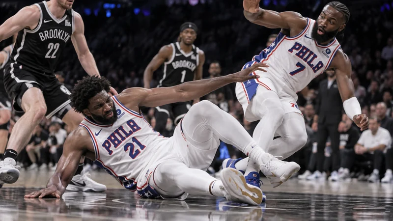 Realistic court-level Sixers game scene with Joel Embiid in white No. 21 on the floor after a scary collision as a teammate reacts nearby.