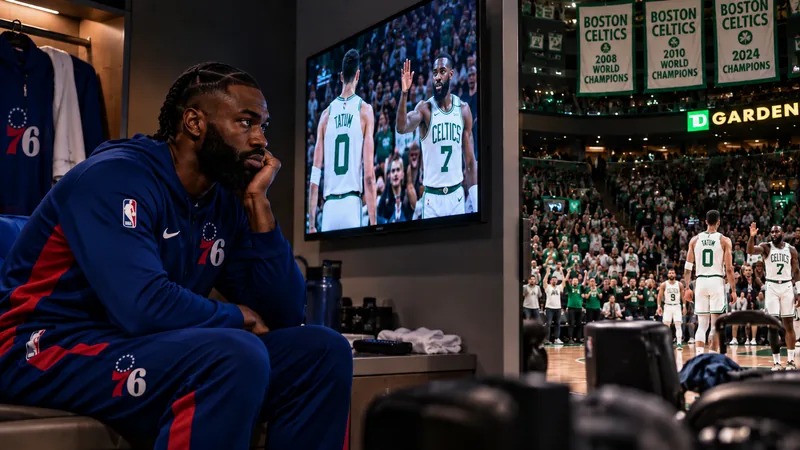 Satirical locker-room scene with a Jaylen Brown-like player in current Philadelphia 76ers gear sitting alone and watching a Celtics game at TD Garden.