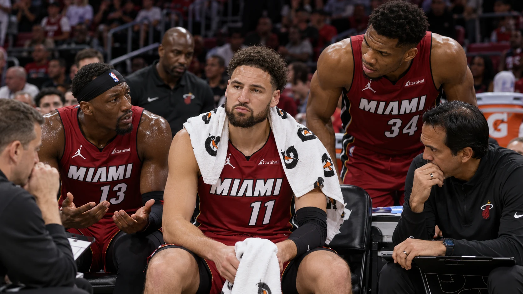 Klay Thompson sitting on the Miami Heat bench while Bam Adebayo and Giannis Antetokounmpo lean in during a serious timeout discussion.