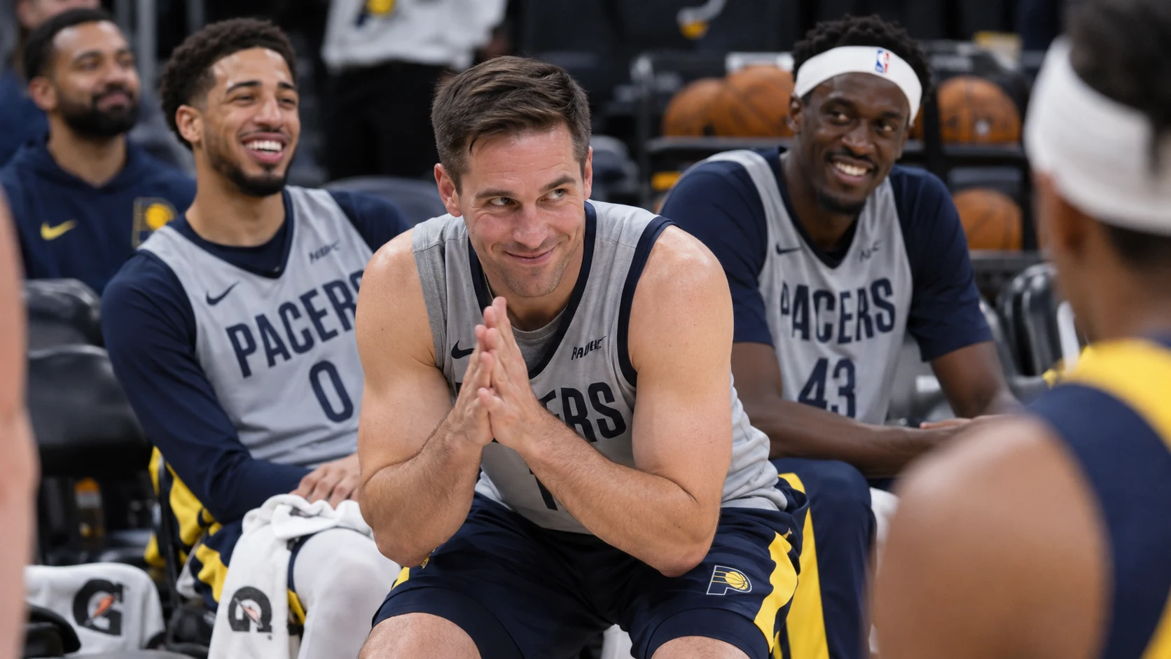 T.J. McConnell crouches on the Pacers bench smiling mischievously while Tyrese Haliburton and Pascal Siakam sit behind him.